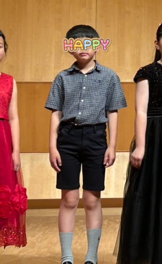 Young boy in a blue-gray checkered shirt and black shorts standing on a wooden stage, face covered by a colorful 'HAPPY' sticker.