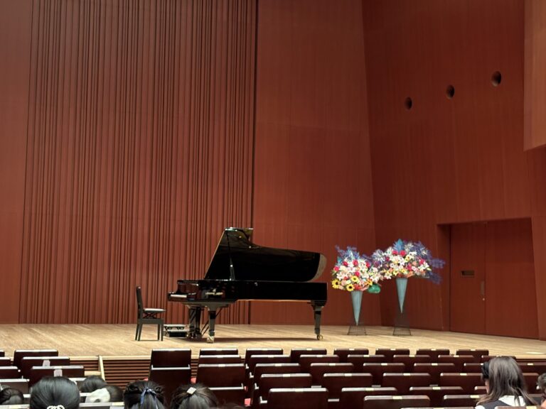 Stage with a grand piano and flower arrangements on a wooden-panel backdrop, audience seating in the foreground.